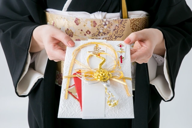 A close-up of a woman in traditional Japanese attire holding an ornate Shūgi-bukuro envelope decorated with gold mizuhiki knots, symbolizing celebration and good fortune, typically used for monetary gifts at weddings and other formal events.
