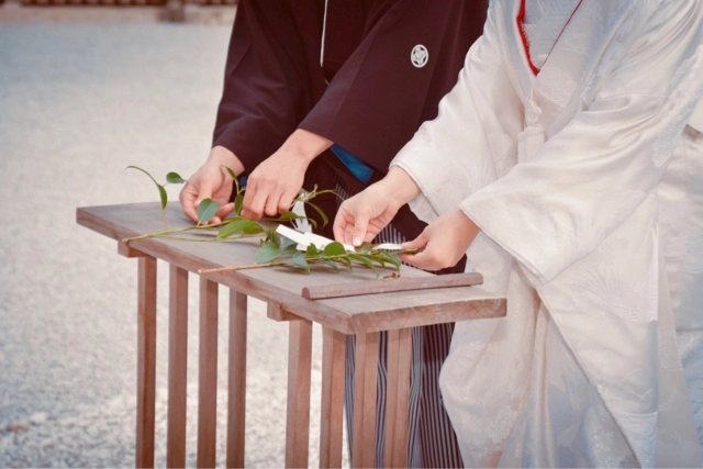 Bride and groom dressed in traditional Shinto wedding attire performing a sacred ritual offering with greenery at a ceremonial altar.