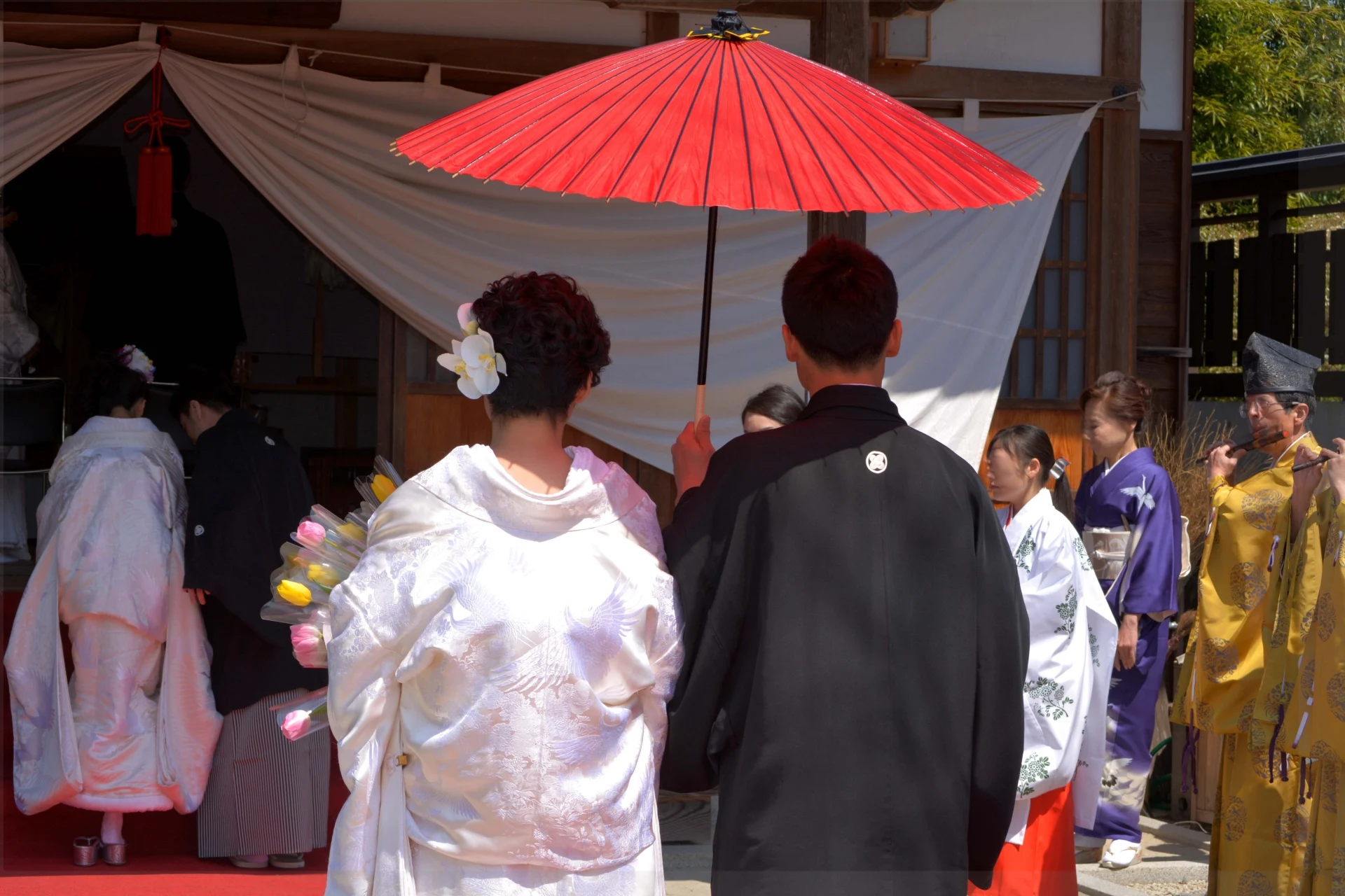A Shinto wedding ceremony scene featuring the bride in a white kimono and the groom in a black montsuki, holding a red parasol, surrounded by attendants in traditional Japanese attire.
