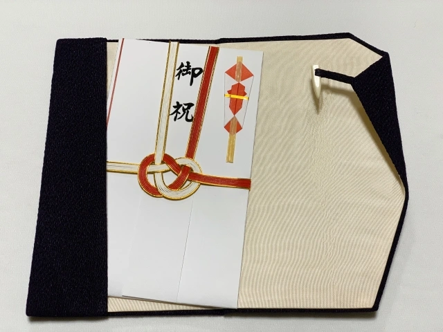 A traditional Japanese Shūgi-bukuro envelope adorned with a red and gold mizuhiki knot, placed inside a protective fabric cover, used for presenting monetary gifts during celebratory occasions like weddings or anniversaries.