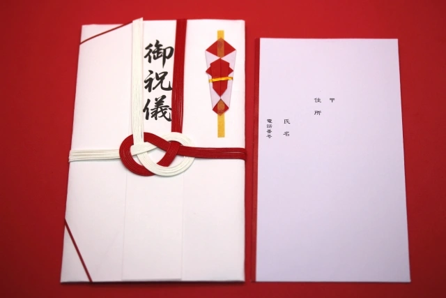 A traditional Japanese Shūgi-bukuro envelope decorated with a red and white mizuhiki knot and kanji characters, accompanied by an inner slip for writing the recipient's name, used for monetary gifts during celebratory occasions like weddings or anniversaries.