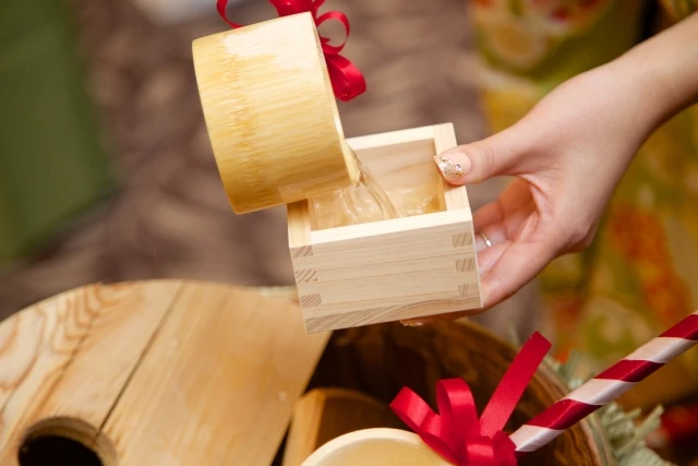 A hand pouring sake from a bamboo ladle into a wooden masu cup during the Kagamibiraki ceremony, highlighting the traditional Japanese serving ritual.