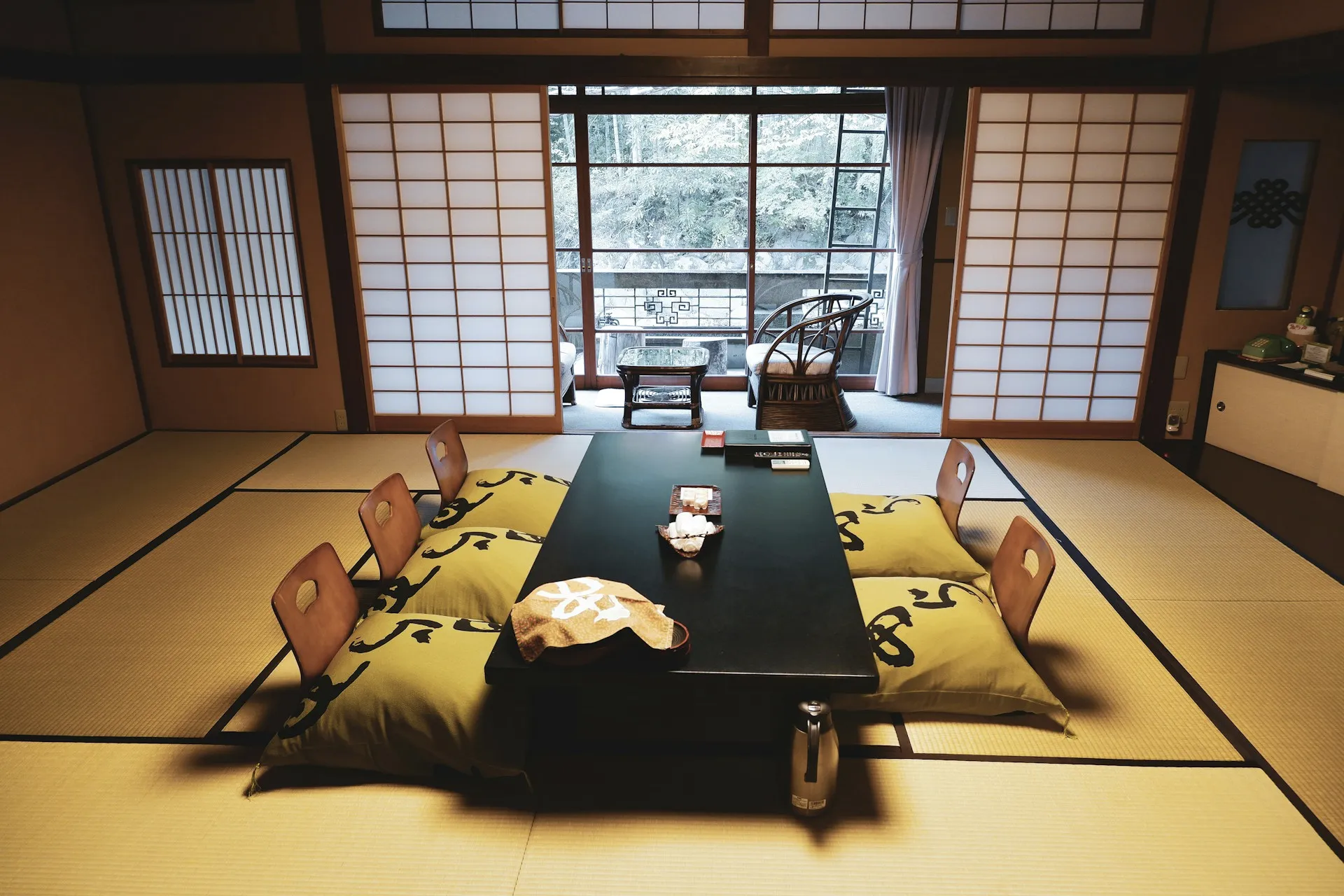 Interior of a Japanese tatami room with floor cushions, a low table, and sliding shoji doors opening to a garden view.