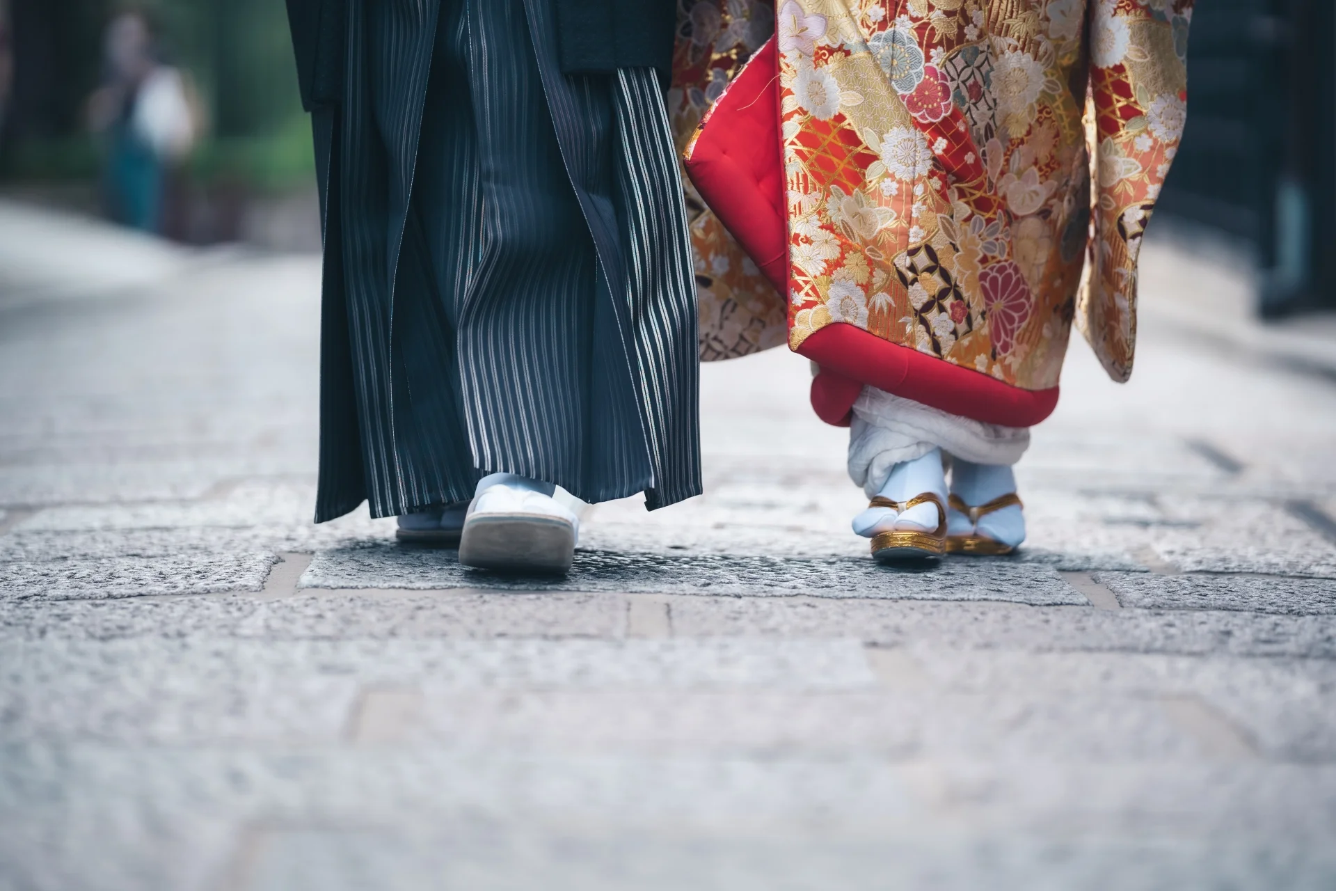 Close-up of a couple wearing traditional Japanese wedding attire, with the groom in striped hakama and the bride in an ornate kimono, walking side by side on a stone-paved street.