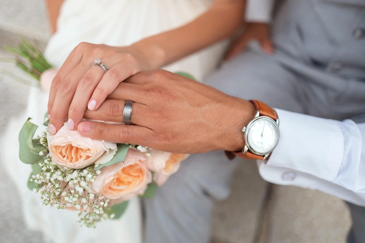 Close-up of a bride and groom's hands with wedding rings resting on a bouquet of pale peach roses, showcasing their wedding bands and engagement ring.