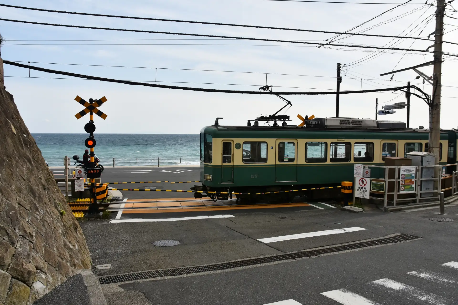 A green and yellow tram is stopped at a level crossing with a red signal light, near Kamakura coastal road with the ocean in the background.