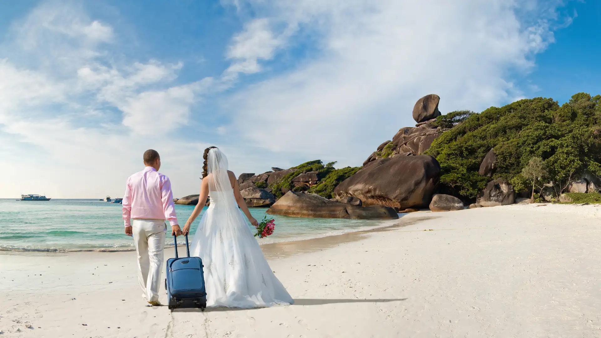 Newlywed couple in wedding attire walking hand-in-hand along a pristine tropical beach with turquoise water and granite boulders.