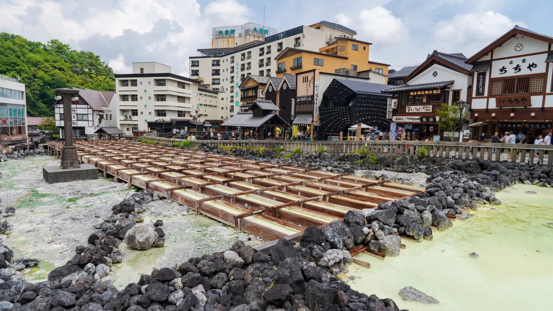 Yubatake hot spring source at Kusatsu Onsen, wooden cooling frames over milky-green water surrounded by traditional ryokan buildings.