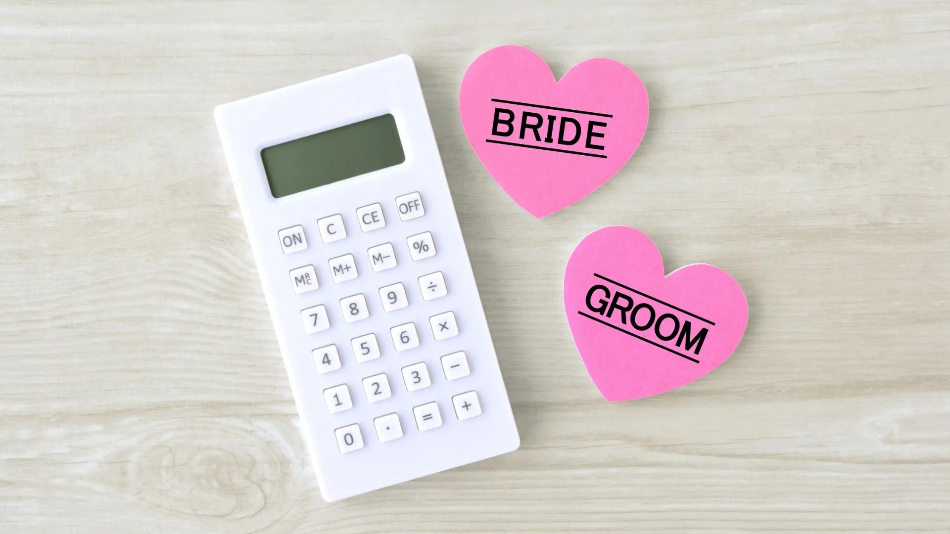 A white calculator beside two pink heart-shaped notes labelled Bride and Groom on a wooden surface, representing budget planning for a Japan destination wedding
