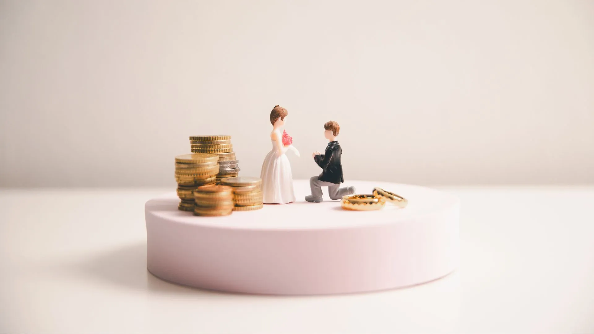 Miniature bride and groom figurines standing beside stacked coins and gold rings on a pink platform, symbolising the cost of planning a destination wedding in Japan