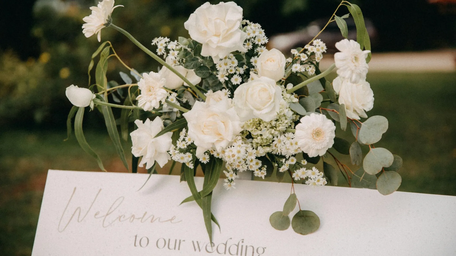 White floral arrangement with roses and eucalyptus on a welcome sign that reads “Welcome to our wedding” for a Japanese wedding reception