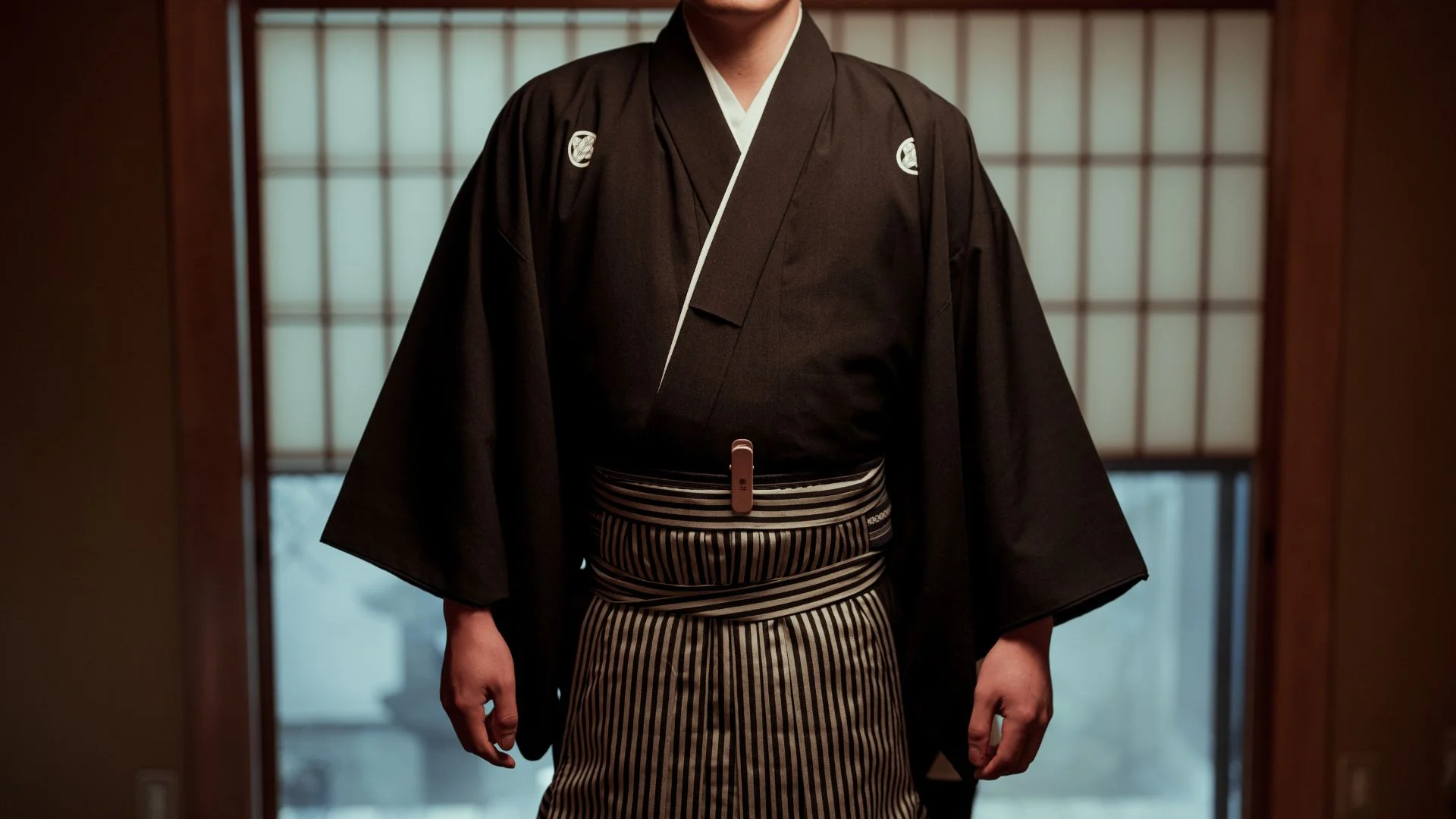 Person wearing a black kimono with white collar lining and striped hakama, standing indoors at the doorway area.