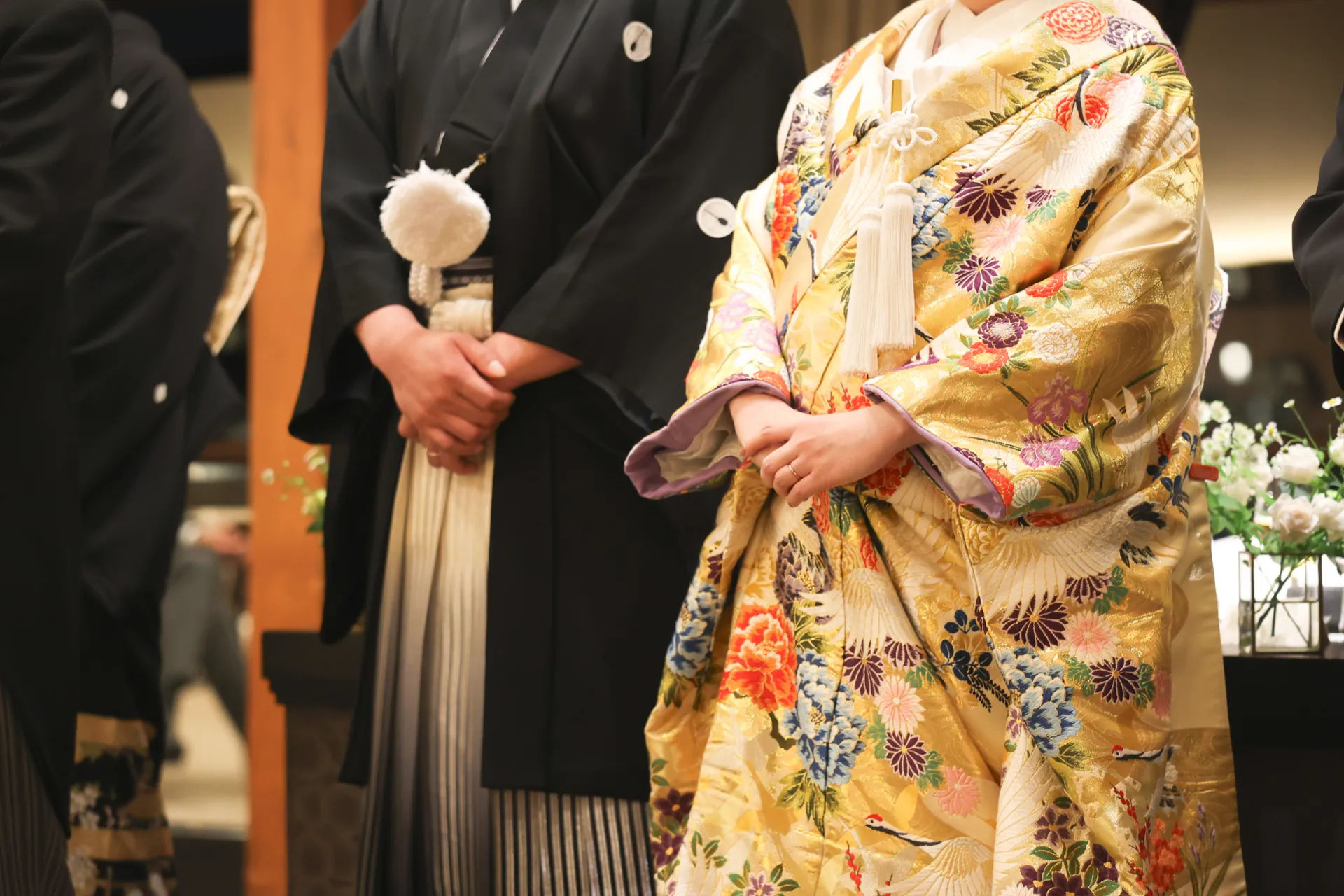 Bride wearing elegant gold iro uchikake with colorful floral and crane embroidery, standing with groom in traditional black attire.