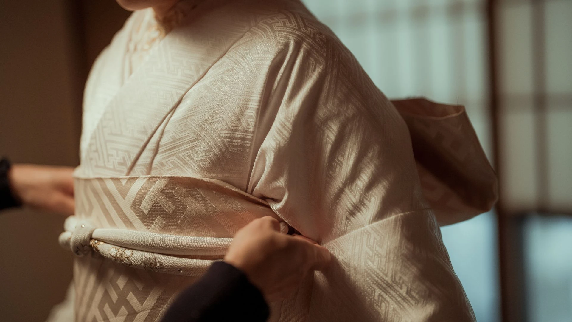 Close-up of a traditional Japanese shiromuku wedding kimono being carefully tied with a white obi sash, showing intricate geometric pattern and obi cord detail
