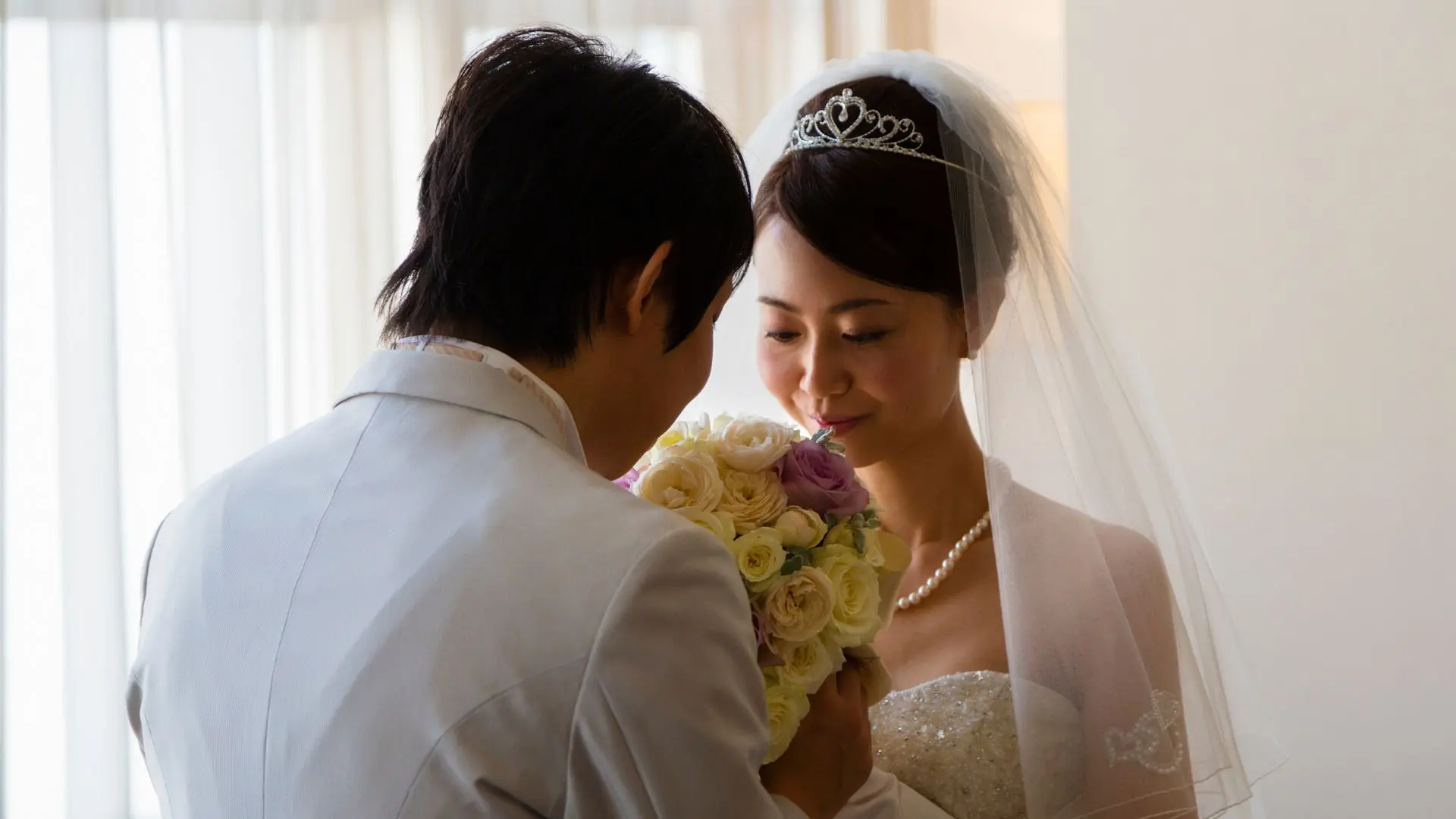 Japanese bride and groom sharing an intimate moment during their Western-style wedding ceremony. The bride wears a sparkling tiara, veil, and white wedding dress while holding a beautiful flower bouquet.