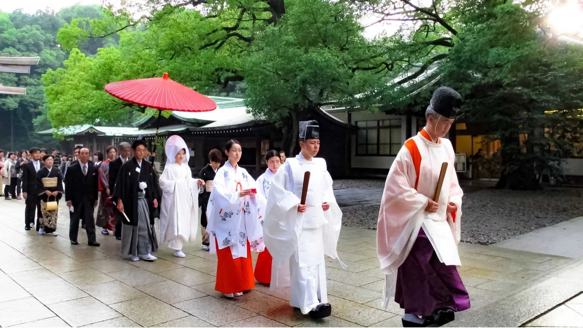 Traditional Shinto wedding procession at a Japanese shrine. The bride wears a white shiromuku kimono with wataboshi hood, accompanied by the groom and Shinto priests in ceremonial robes, walking under a red wagasa umbrella.