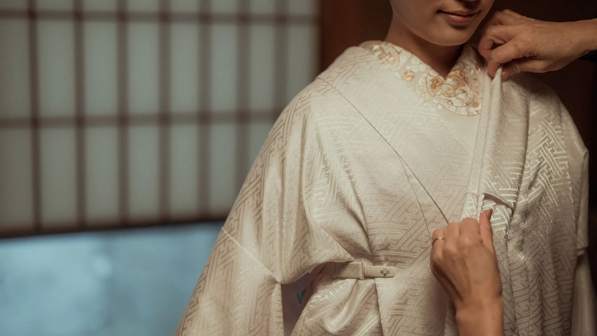 Close-up of hands adjusting the white eri collar on a traditional Japanese shiromuku wedding kimono, showing intricate gold embroidery and proper dressing technique